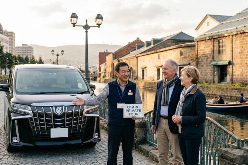 A car with a sign reading 'OTARU PRIVATE TOUR' in front of a canal with buildings in the background. 1