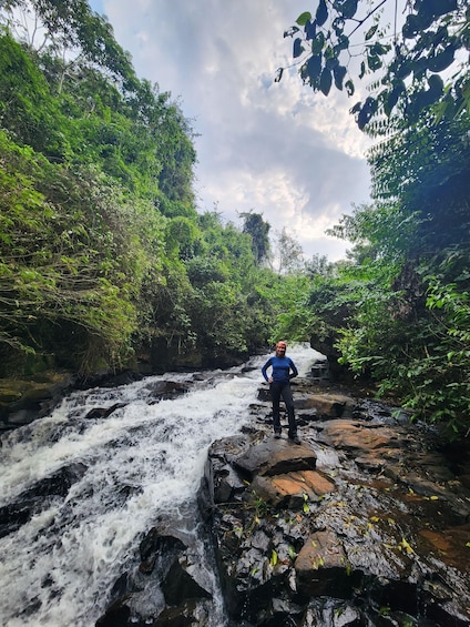 A person standing on rocks by a flowing river surrounded by dense greenery. 2