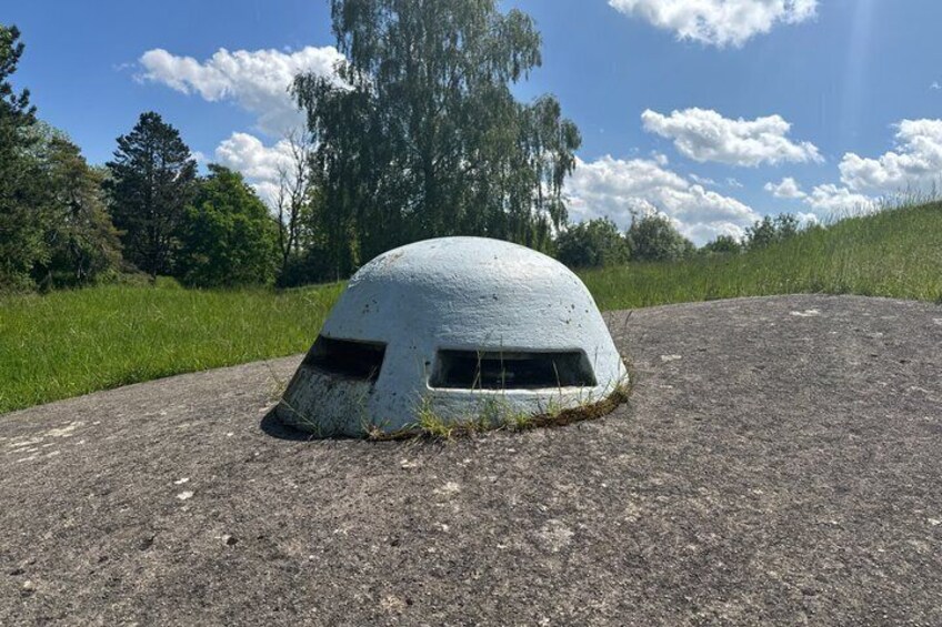 A concrete bunker-like structure on a gravel surface with a grassy field and trees in the background. 3