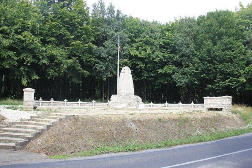 A memorial monument with a flagpole, surrounded by a white fence and steps leading up to it, set against a backdrop of dense forest. 1