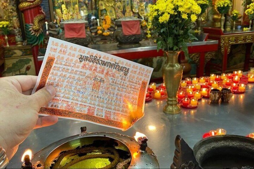 A hand holding a paper with text, a lit candle, and a vase with yellow flowers in a temple setting. 5