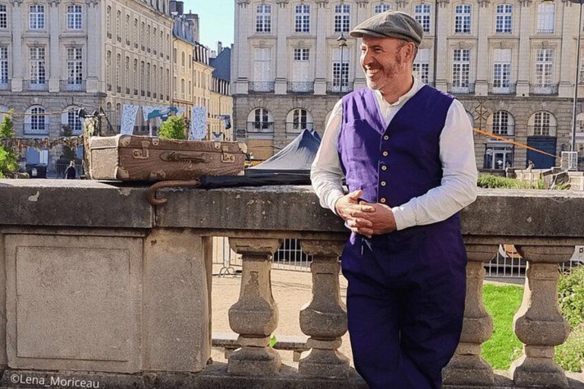 A man standing on a stone balustrade with a suitcase and a hat, overlooking a historic building with classical architecture. 5