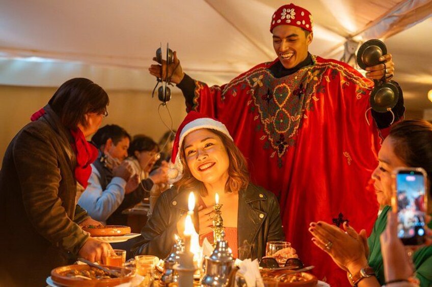 A person in traditional attire holding a camera, a lit candle, and a glass of drink at a festive gathering. 5