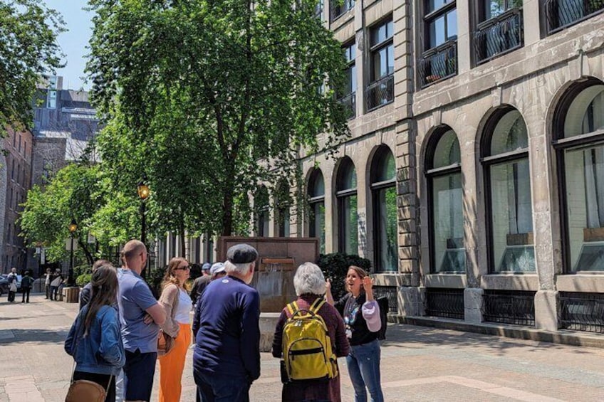 A group of people standing on a city sidewalk, facing a building with arched windows and a stone facade. 2