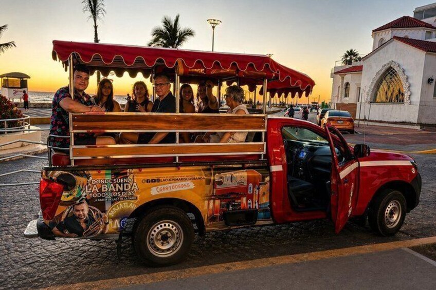 A red truck with a covered back, advertising 'Donde Suena La Banda,' parked on a cobblestone street with a sunset in the background. 1