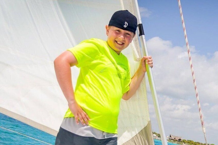 A person in a neon yellow shirt and dark cap leaning on a sailboat's mast. 5