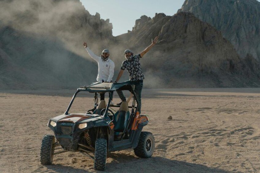 Two people on a dune buggy in a desert with rocky mountains in the background. 5
