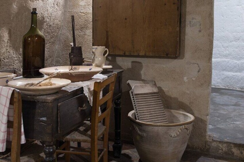 A rustic kitchen with a wooden table, a ceramic jug, a metal grater, and a large clay pot. 3