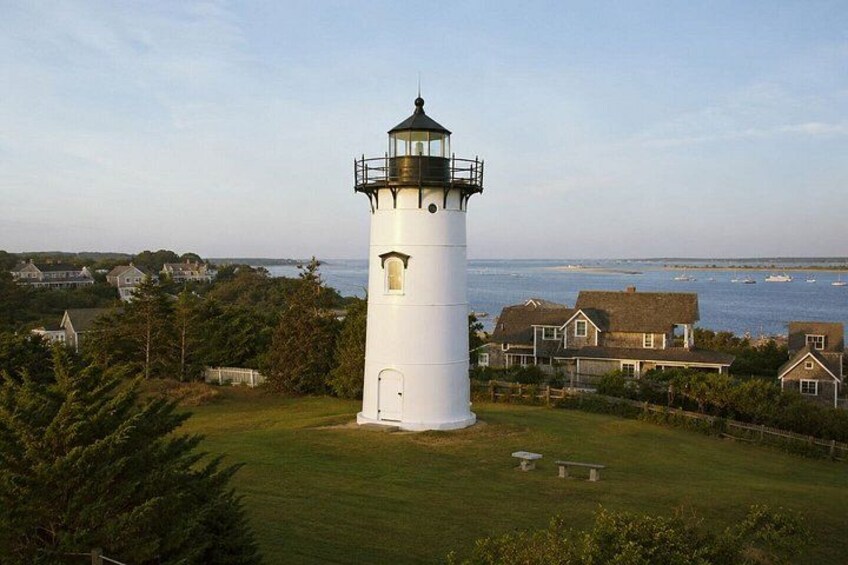 A white lighthouse with a black top, surrounded by houses and trees, overlooking a body of water. 2