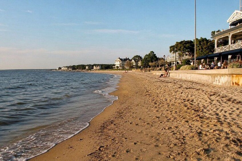 A sandy beach with gentle waves, a promenade with people, and buildings in the background. 3