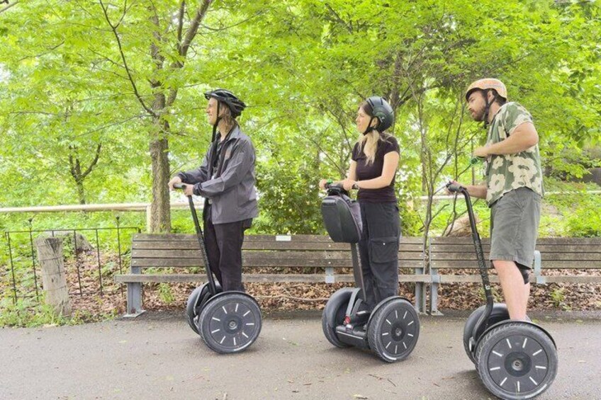 Three people on segways in a park with trees and a bench. 1