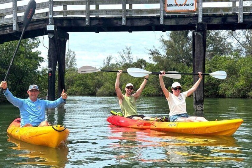Three people kayaking under a bridge with a 'MINIMUM WAKE' sign. 1