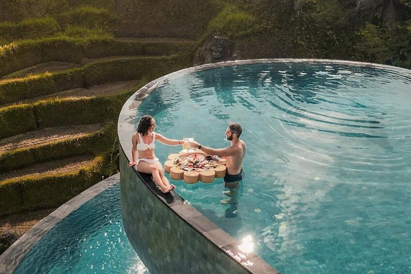 A couple sitting by a pool with a table of drinks and snacks. 1