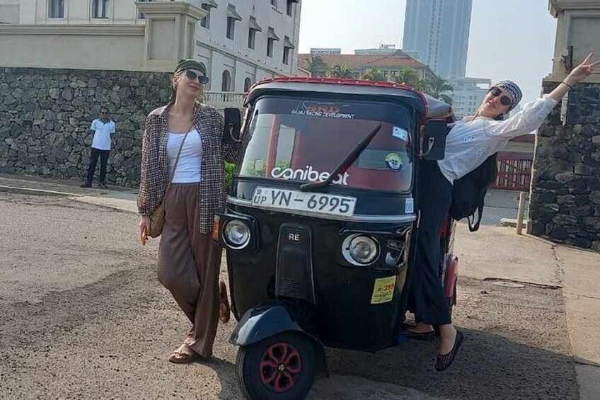 A tuk-tuk parked on a street with a license plate, a person standing next to it, and another person leaning on the vehicle. 1