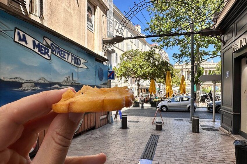 Una mano che tiene un boccone di cibo fritto, una strada con edifici e un cartello con la scritta 'LA MER STREET FOOD'. 2