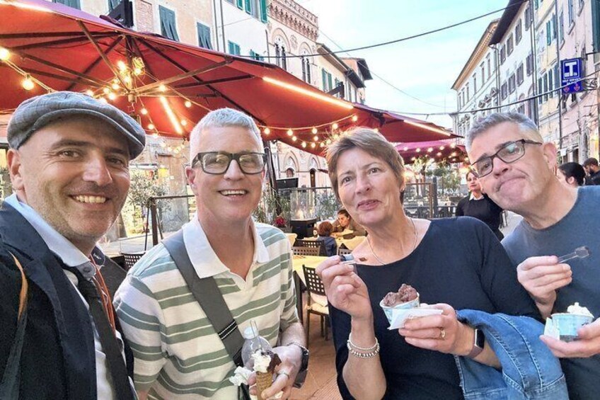 Four people outdoors, holding ice cream cones, with a street and buildings in the background. 1