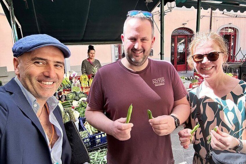 Three people holding green vegetables, possibly cucumbers, in an outdoor market setting with a building in the background. 2