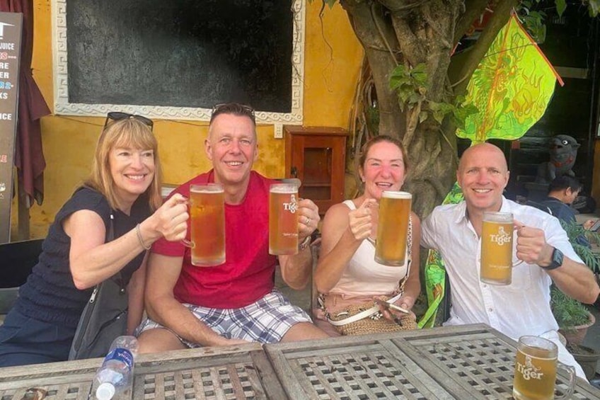 Four people toasting with beer mugs at an outdoor table with a tree and a yellow wall in the background. 2