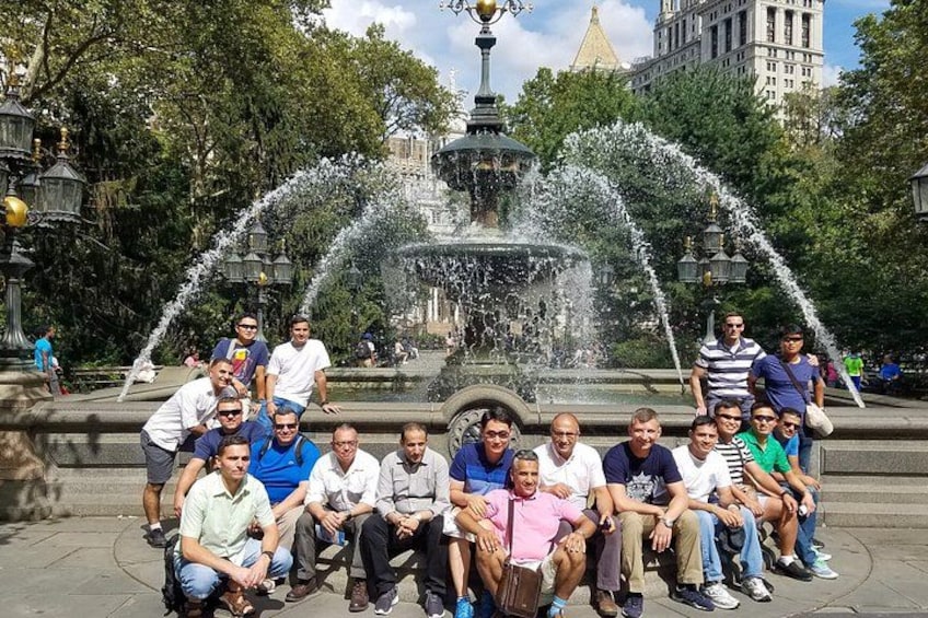 A group of people sitting in front of a fountain with water jets in a park with trees and buildings in the background. 2