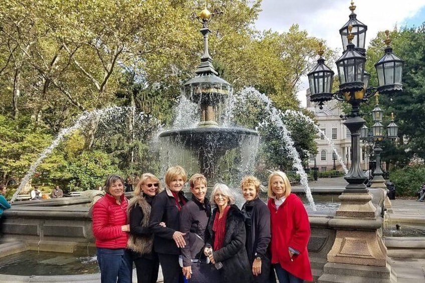 A group of people standing in front of a fountain with water jets, surrounded by trees and historic buildings. 4