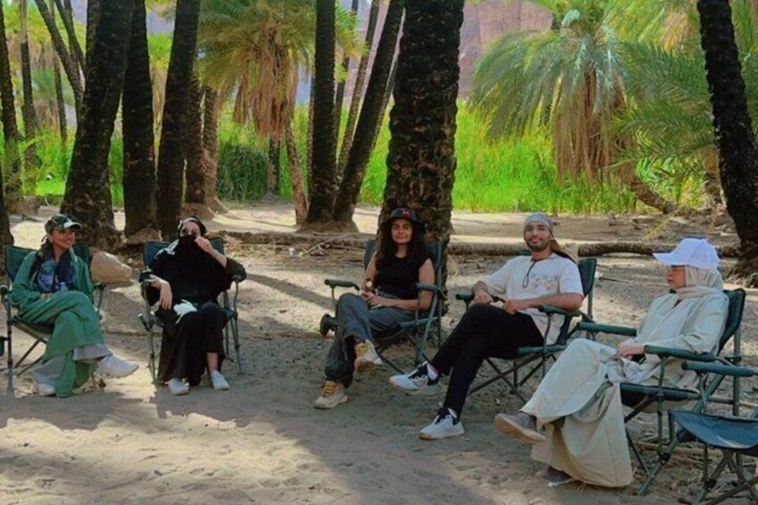A group of people sitting on chairs in a sandy area with palm trees in the background. 5