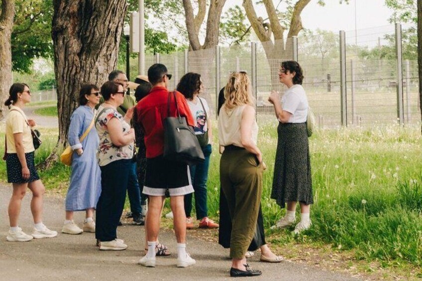 A group of people standing on a pathway in a park with trees and a fence in the background. 4