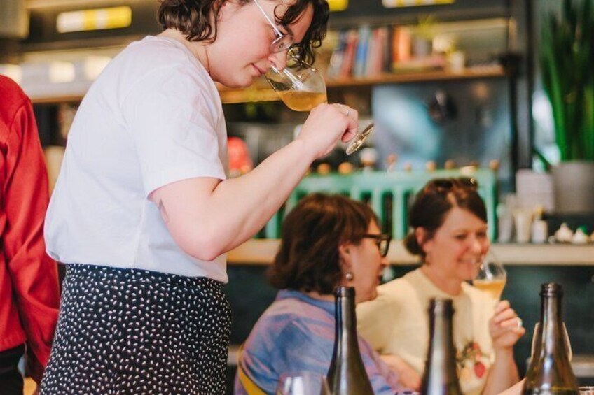 A person pouring a drink into a glass, with bottles on the table and a blurred background of a restaurant setting. 1