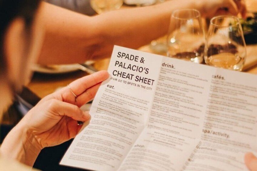 A person holding a menu with the title 'SPADE & PALACIO'S CHEAT SHEET' and sections for 'eat,' 'drink,' 'cafés,' and 'spa/activity.' 5