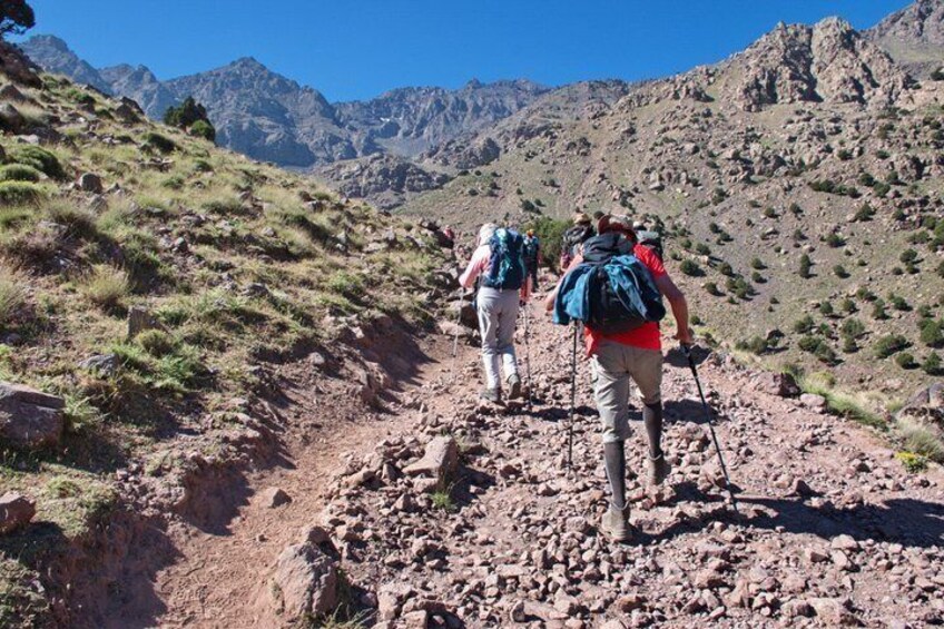 Hikers on a rocky mountain trail with sparse vegetation and distant mountains. 3