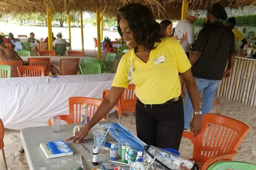 A person in a yellow shirt standing at a table with art supplies, a book, and a plastic chair. 1