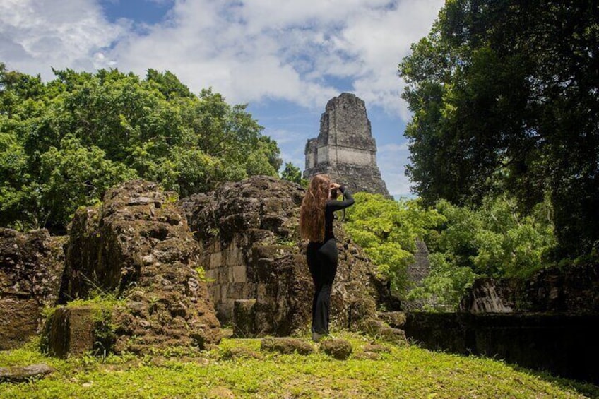 A person taking a photo of a pyramid-like structure amidst ancient ruins and lush greenery. 3