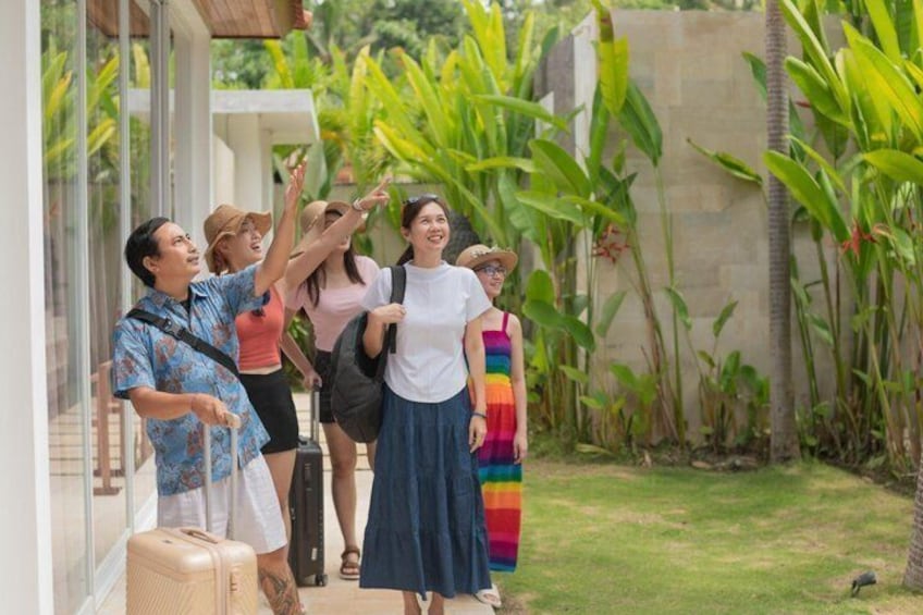 A group of people standing outside a building with a suitcase, tropical plants, and a clear sky. 3