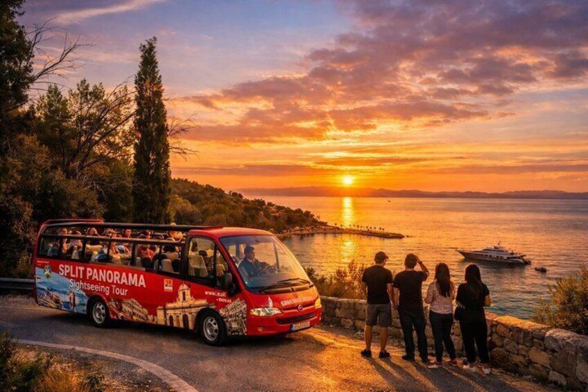 A red tour bus with 'SPLIT PANORAMA Sightseeing Tour' on its side, parked by a waterfront with a sunset in the background. 1