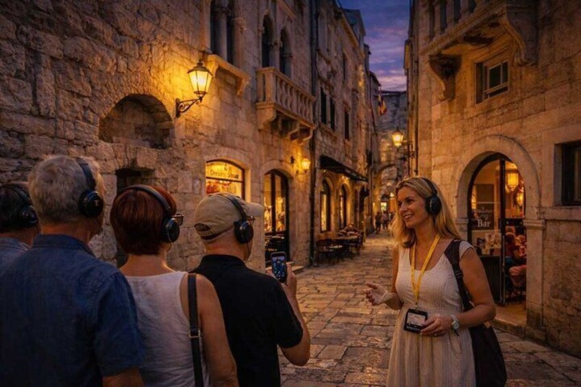 A group of people walking down a cobblestone street at dusk, with buildings on either side and street lamps providing light. 5