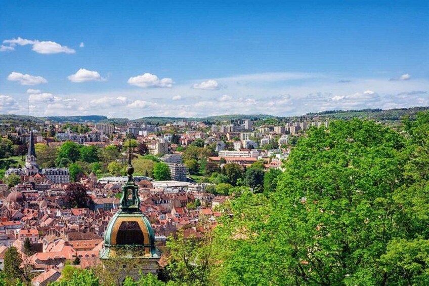 En stadsbild med en framträdande byggnad som har ett stort, grönt kupoltak, täta byggnader och en klarblå himmel. 3