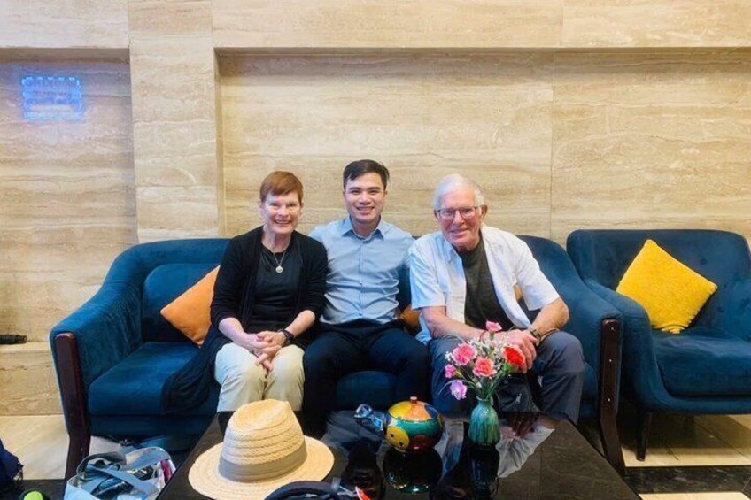 Three people seated on a blue couch with a yellow pillow, a hat, and a vase with flowers on a table. 3