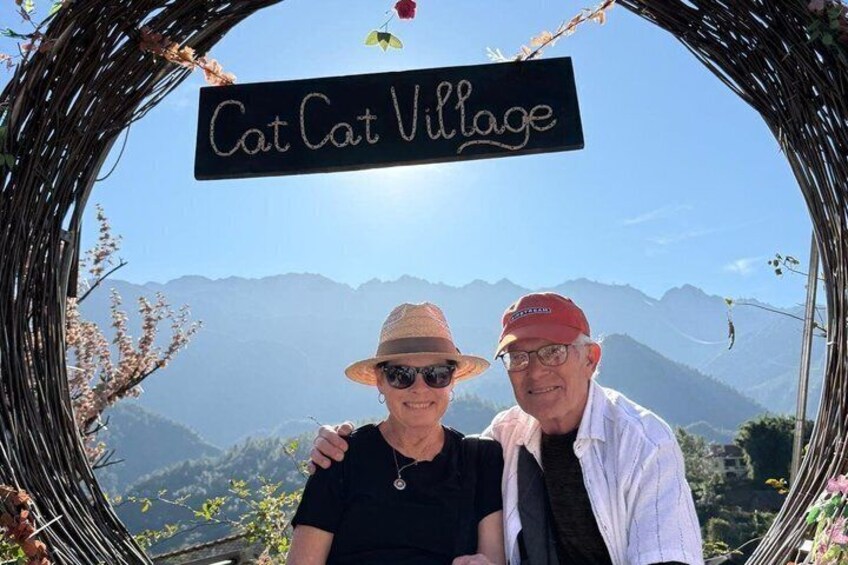 A couple posing in front of a sign that reads 'Cat Cat Village' with a mountainous landscape in the background. 1