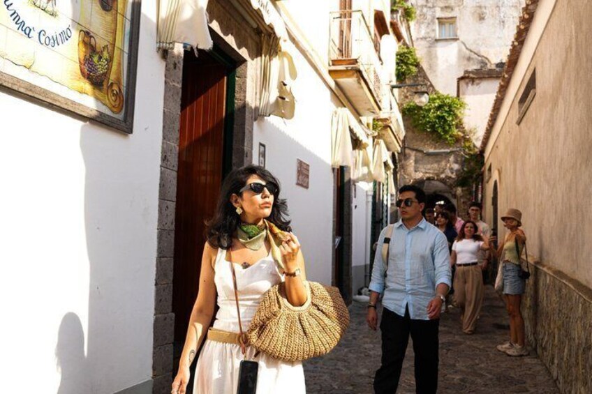 A narrow street with buildings, a person walking with a straw bag, and others in the background. 4