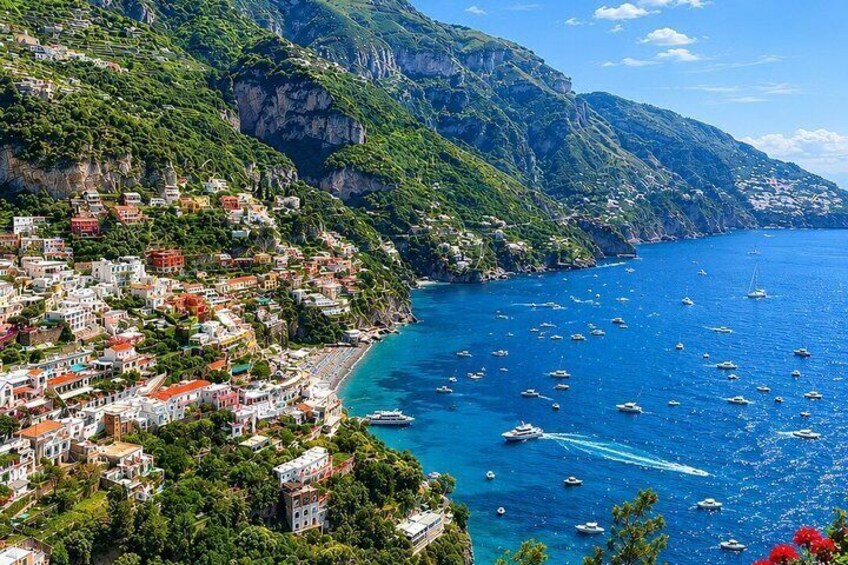 A coastal town with buildings on a hillside, a clear blue sea, and boats. 1