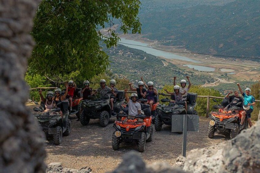 A group of people on quad bikes, a dirt path, and a scenic landscape with a river and hills in the background. 3