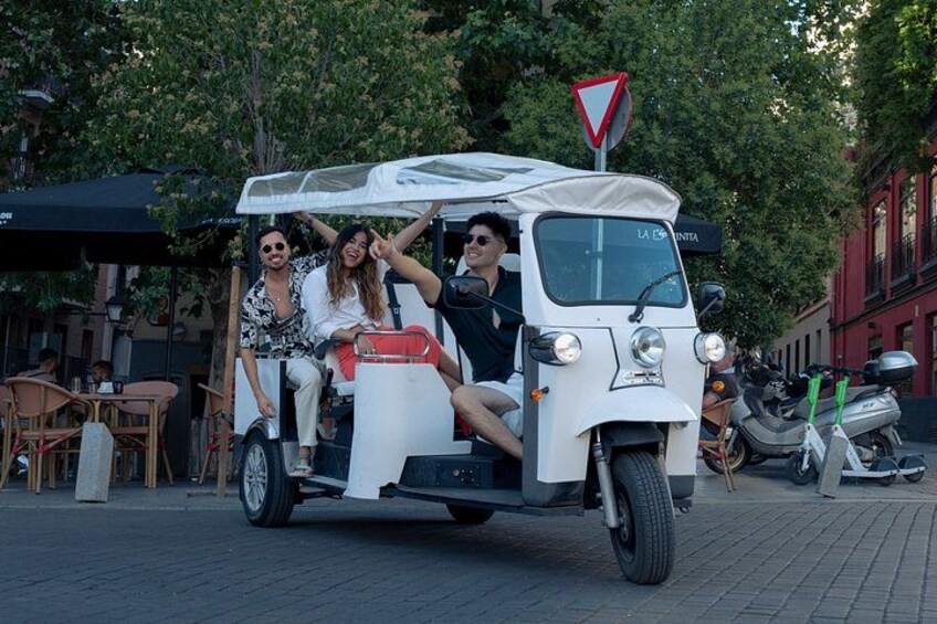 A three-wheeled vehicle with passengers, parked on a street with trees and buildings in the background. 3