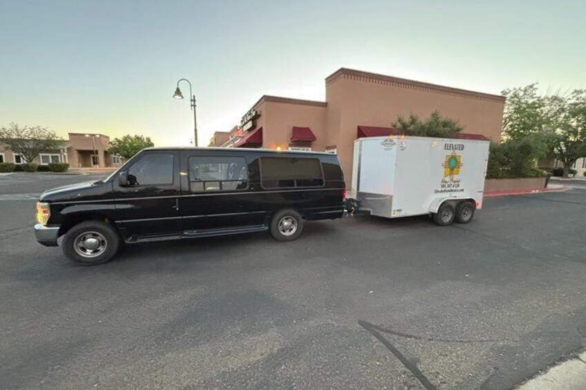 A black van towing a white trailer with 'ELEVATED' branding, parked in a lot with buildings in the background. 2