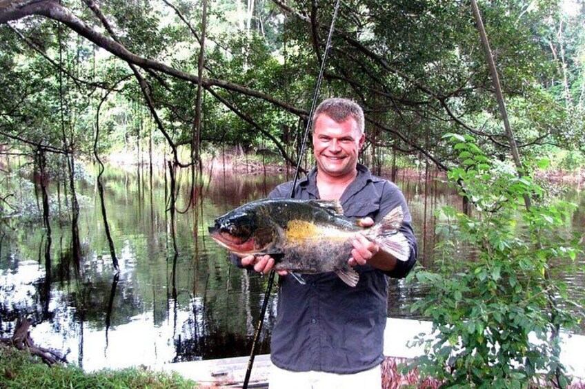 A person holding a large fish in a forested area with a body of water. 3