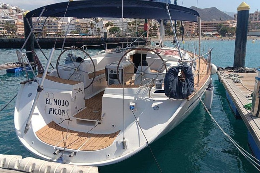 Een witte boot, genaamd 'El Mojo Picón', afgemeerd in een jachthaven met een houten dek en een luifel. 1