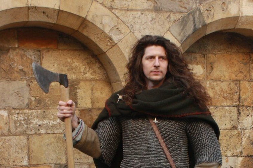 A person holding an axe, wearing a chainmail shirt and a cloak, standing in front of a stone building with arches. 2
