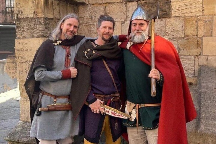 Three people in medieval costumes standing in front of a stone building. 1