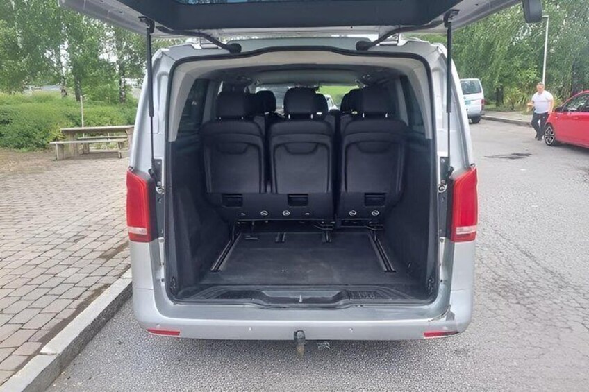 A silver van with its rear cargo area open, revealing a flat loading surface and two sets of black seats. 2