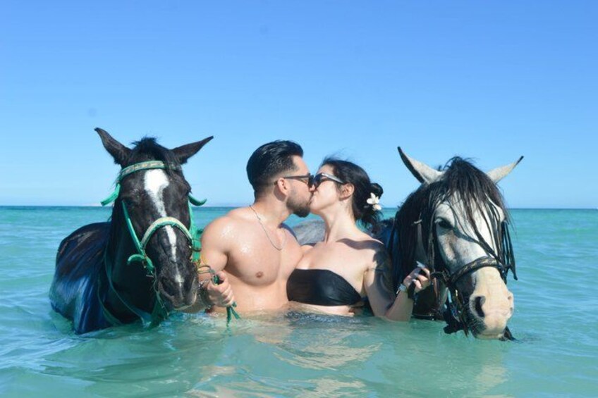 Una pareja en aguas poco profundas con dos caballos, uno negro y uno blanco, en un mar azul cristalino. 2