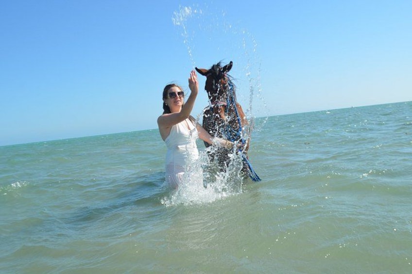 A woman in a white dress splashing water on a horse in the ocean. 1