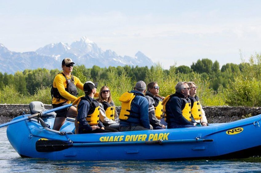 A group of people in a blue inflatable raft labeled 'Snake River Park' on a body of water with mountains in the background. 2
